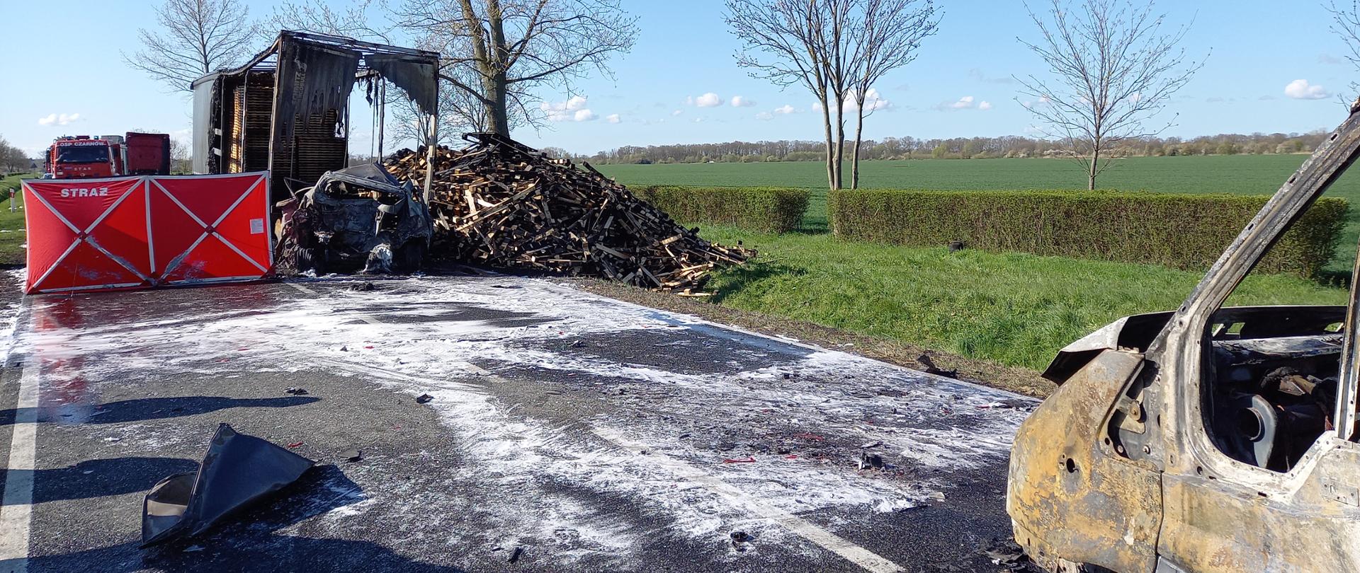 Z prawej strony przód spalonego samochodu dostawczego. Dalej widoczny spalony wrak samochodu osobowego wbity pod tył naczepy samochodu ciężarowego. Z prawej strony naczepy w rowie i na poboczu drogi sterta pozostałości po paletach przewożonych przez samochód ciężarowy. Z lewej czerwony parawan z napisem Straż.