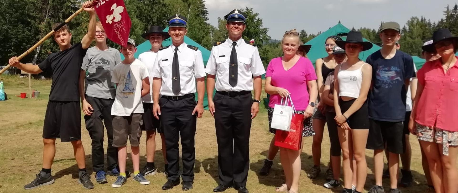 Na zdjęciu widać grupę ludzi znajdujących się na trawniku. W środku znajdują się dwaj funkcjonariusze Państwowej Straży Pożarnej. Obok nich stoją osoby w cywilnym ubraniu trzymający proporzec oraz torebki. Zdjęcie wykonane w dzień na placu obozu harcerskiego. 