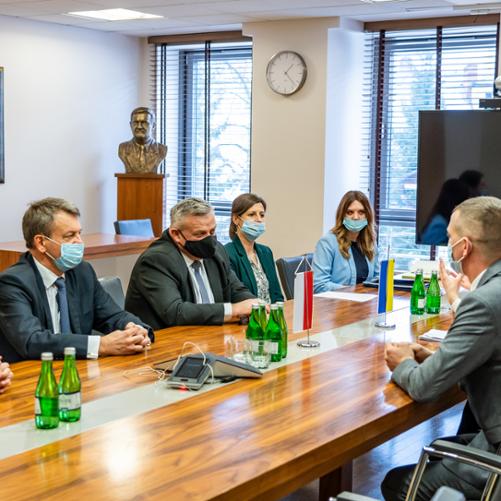 Ludzie siedzący po obydwu stronach stołu. Na stole ustawione są dwie małe flagi Polski i Ukrainy oraz butelki z wodą.