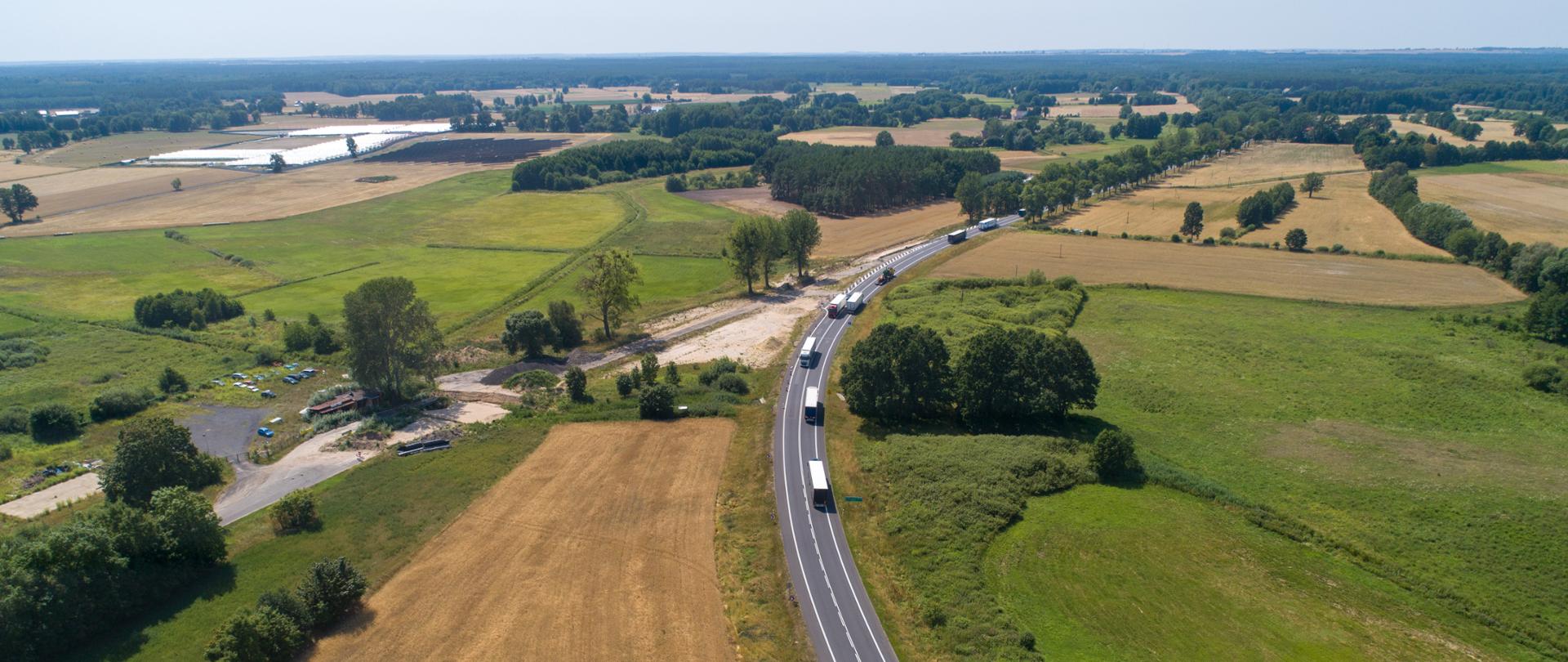 Zdjęcie z lotu ptaka. Droga krajowa otoczona polami i fragmentami lasu, na drodze jadące samochody. Widoczne miejsce z rozpoczętymi pracami budowlanymi.