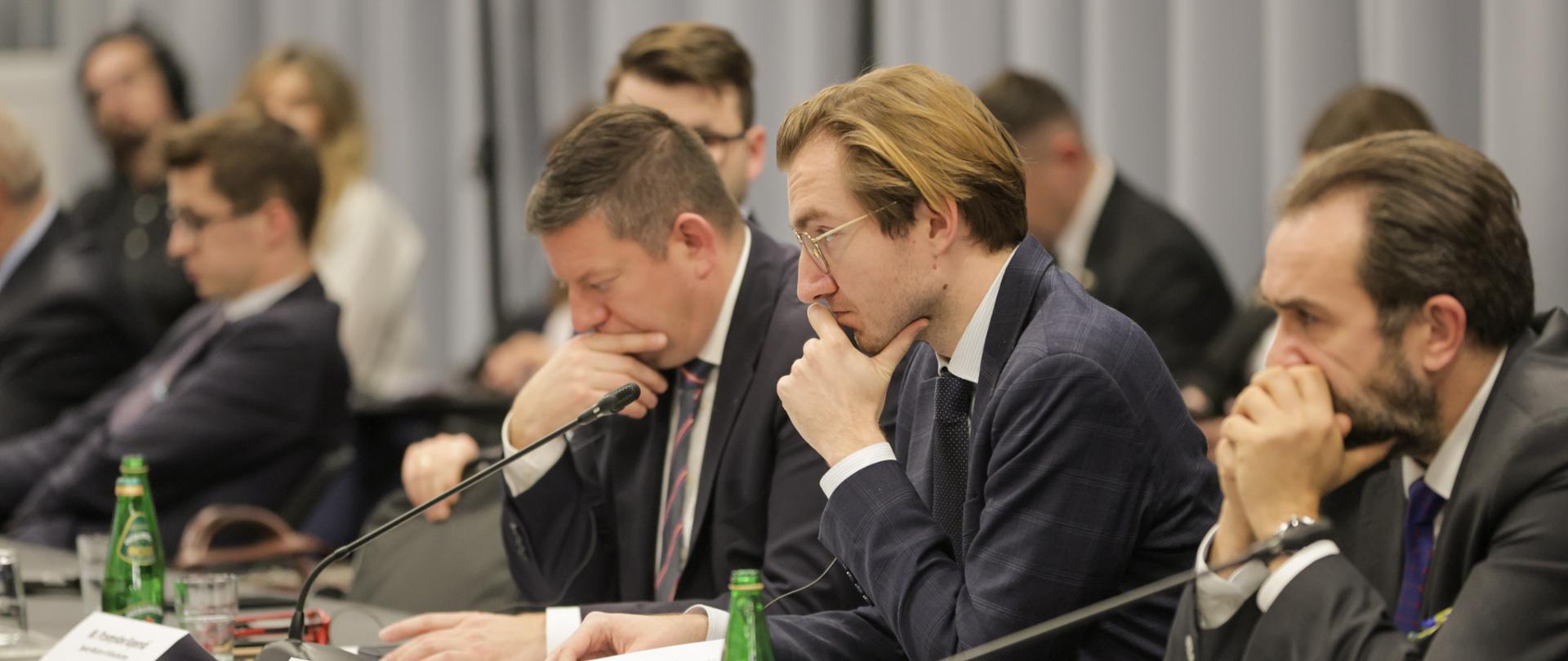 W sali konferencyjnej przy dwóch długich stołach siedzą osoby