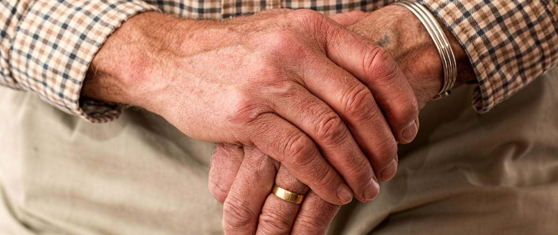 Zdjęcie, na którym widać dłonie starszego mężczyzny trzymającego laskę do podparcia się. Na dłoni mężczyzny widoczna szeroka, złota obrączka. 