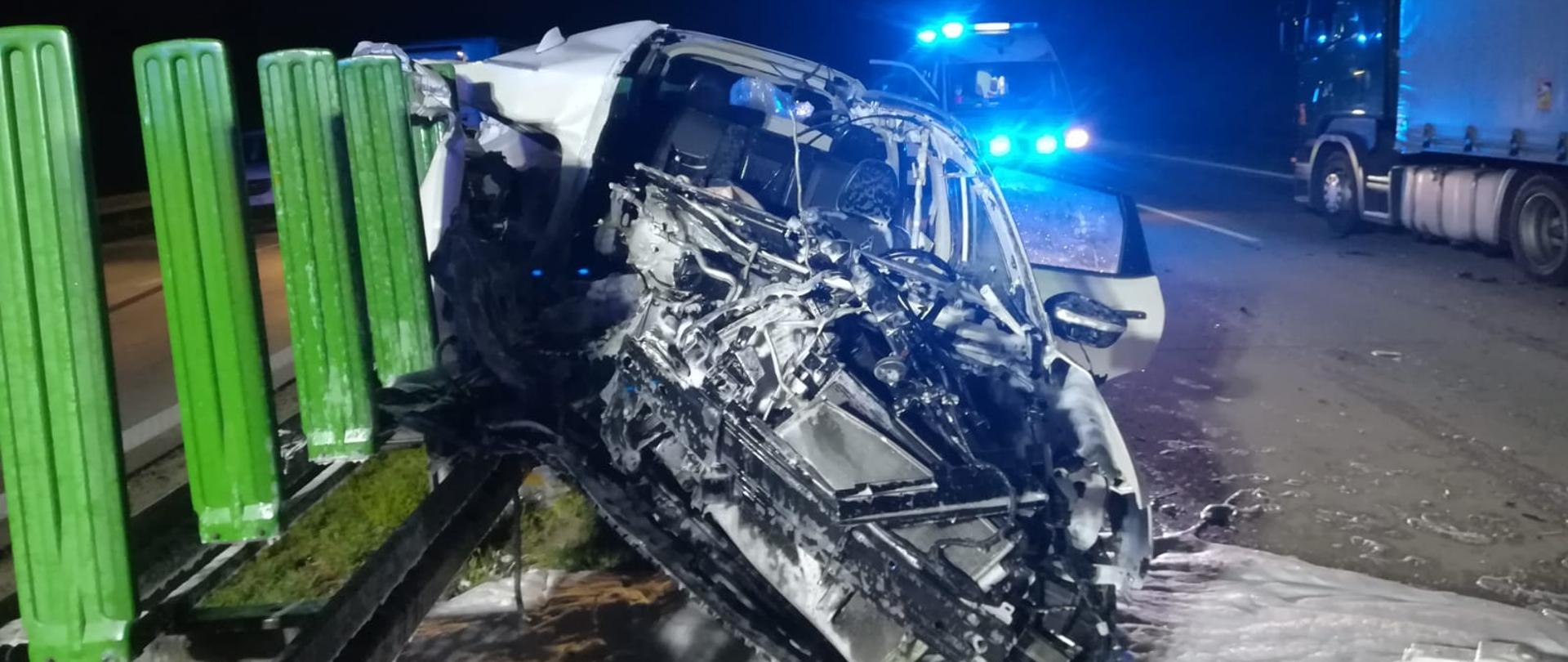 na pierwszym planie rozbity w wyniku wypadku auto osobowe, z tyłu karetka z włączonymi światłami niebieskimi oraz samochód ciężarowy