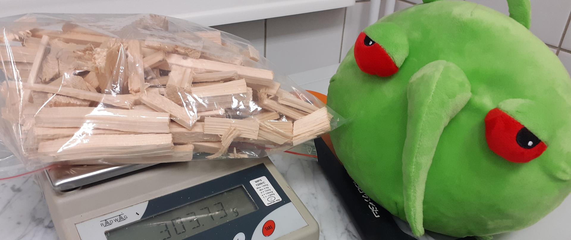 Od lewej waga, na której leży woreczek z fragmentami drewna, na wyświetlaczu 303 g , obok zielona maskotka