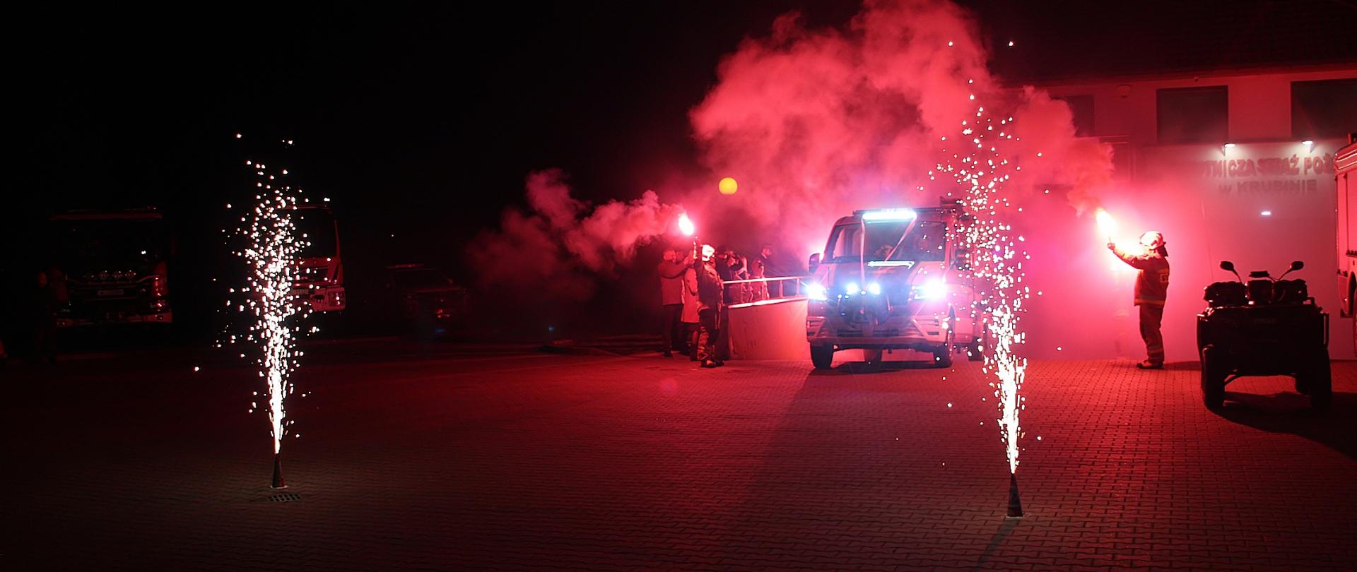 Na plac wjeżdża samochód strażacki między dwiema wysokimi racami. Dookoła samochodu unosi się czerwony dym, przysłaniający czarne niebo. 