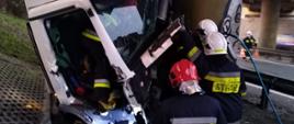 Zdjęcie przedstawia samochód ciężarowy po uderzeniu w filar wiaduktu. Po prawej stronie strażacy wykonujący prace polegające na uwolnieniu osoby poszkodowanej. W wewnątrz kabiny znajduje się strażak nadzorujący parametry życiowe osoby poszkodowanej. 