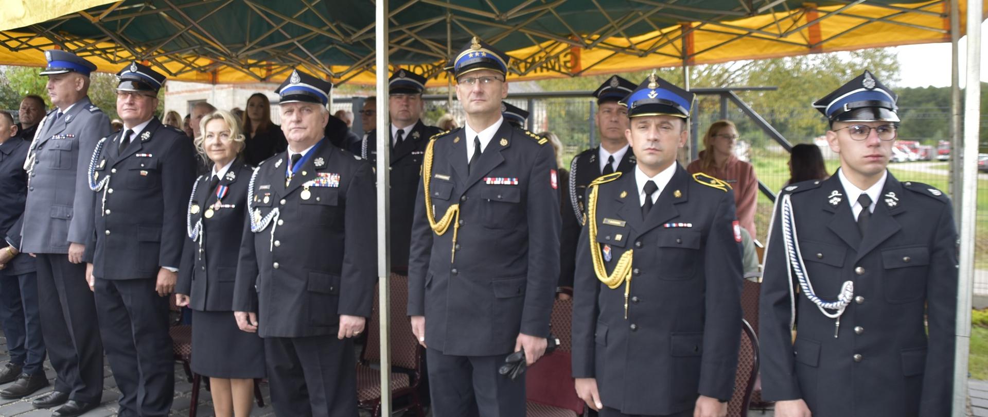 W rzędzie pod namiotem okolicznościowym stoi komendant powiatowy PSP w Żyrardowie st. bryg. Mariusz Tymoszewicz i asp. Tomasz Badowski oraz inni zaproszeni na uroczystość goście honorowi
