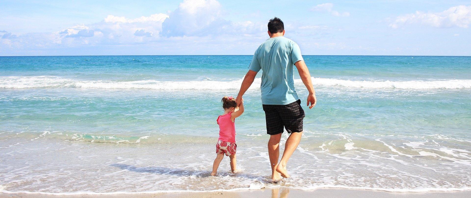Zdjęcie przedstawia plaże nad morzem. W centrum ojciec trzymający córkę za rękę wchodzą do morza.