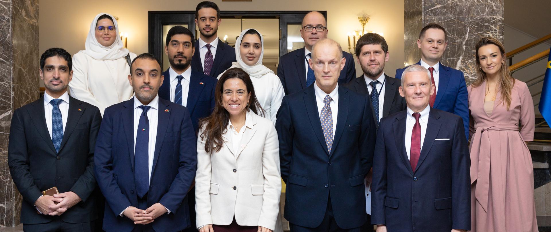 Grupa kilkunastu osób w formalnych strojach pozuje do wspólnego zdjęcia w reprezentacyjnym wnętrzu z marmurowymi kolumnami i schodami. W pierwszym rzędzie stoją kobieta i kilku mężczyzn w garniturach, a pozostali uczestnicy ustawieni są za nimi.
