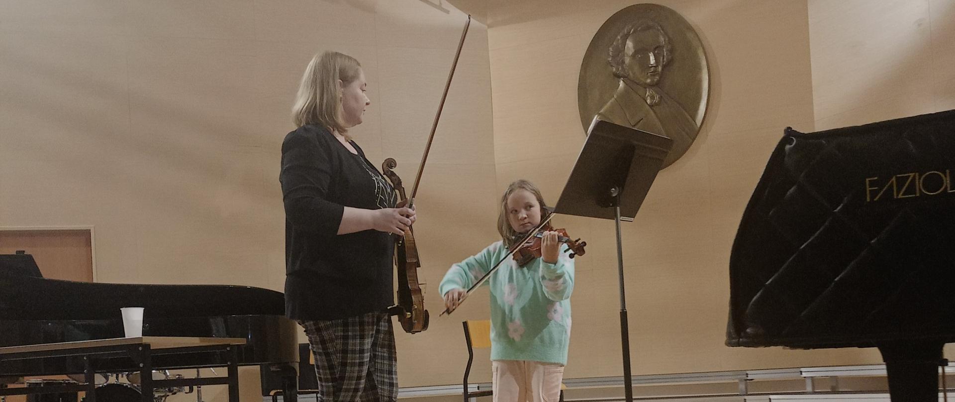 Zdjęcie przedstawia dwie osoby stojące na scenie sali koncertowej. Obie trzymają w dłoniach skrzypce. Na scenie znajduję również dwa fortepiany. 