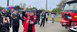 Strażak PSP przemawia przy mównicy podczas deszczowej pogody, osłaniany parasolem przez innego funkcjonariusza. W tle widoczni są uczestnicy uroczystości i flagi Polski.