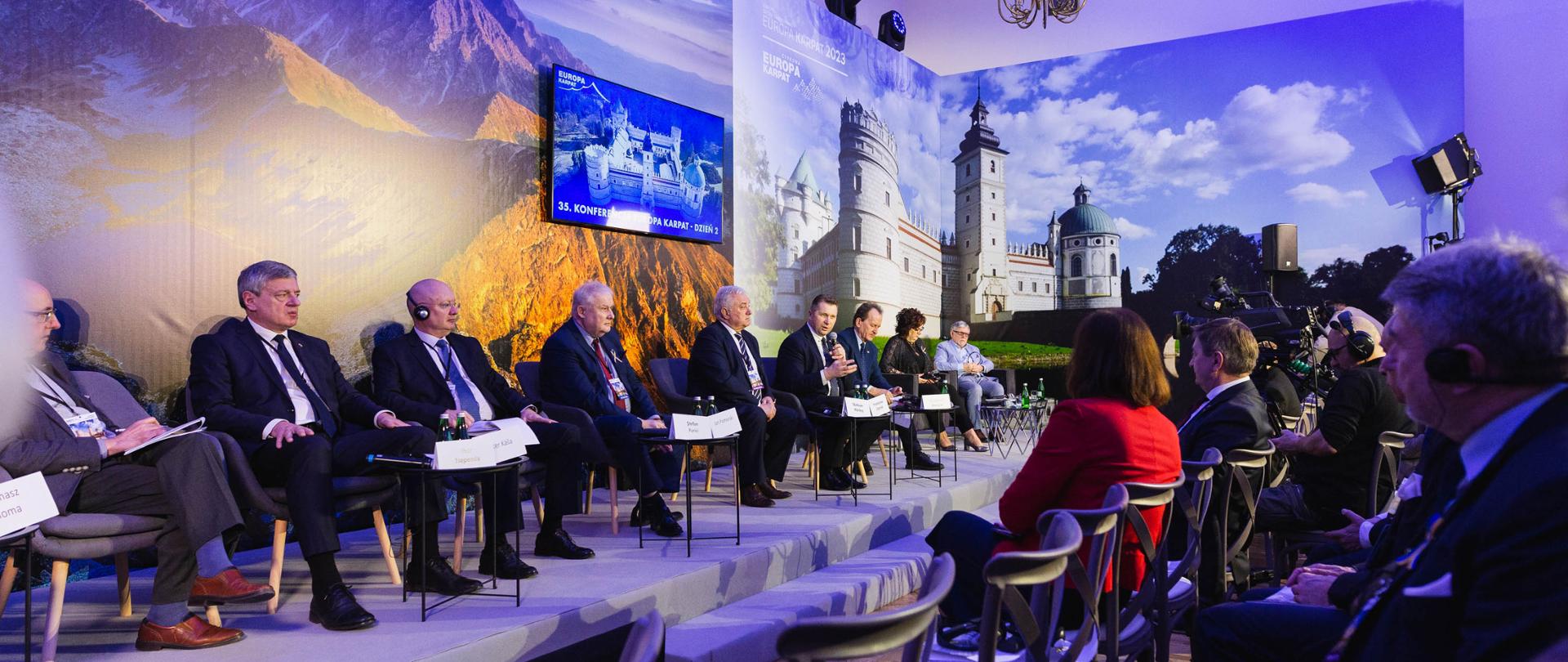 Uczestnicy panelu ośmiu mężczyzn i kobieta siedzą na podeście w fotelach