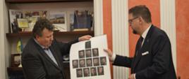 Wiceminister Szeptycki stoi obok mężczyzny w garniturze, razem trzymają dużą kartkę na której są zdjęcia mężczyzn.
