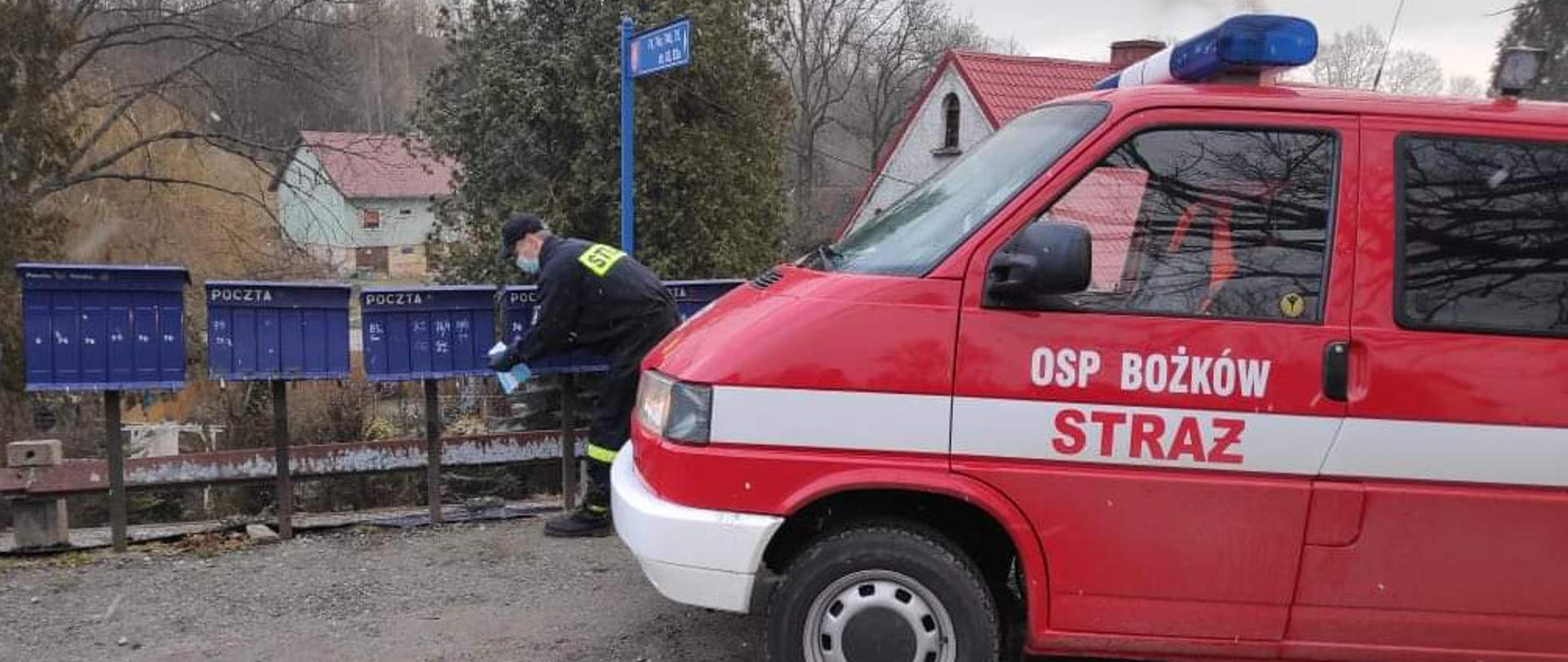 Strażak OSP wrzucający do skrzynek pocztowych ulotki. Po prawej stronie stojący samochód ratownictwa technicznego 