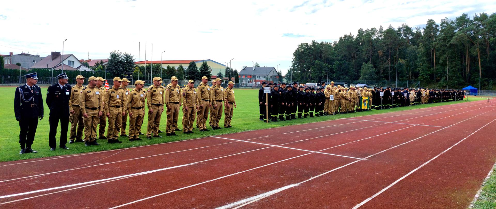 Strażacy PSP i druhowie OSP stoją w równych szeregach na trawiastym boisku, wzdłuż czerwonej bieżni lekkoatletycznej. Część umundurowana jest w czarne galowe mundury, pozostali mają na sobie piaskowe strażackie stroje ćwiczebne z czapkami z daszkiem. W tle widać budynki mieszkalne oraz gęsty pas zielonego lasu pod lekko zachmurzonym niebem. Zbiórka przed zawodami sportowo-pożarniczymi