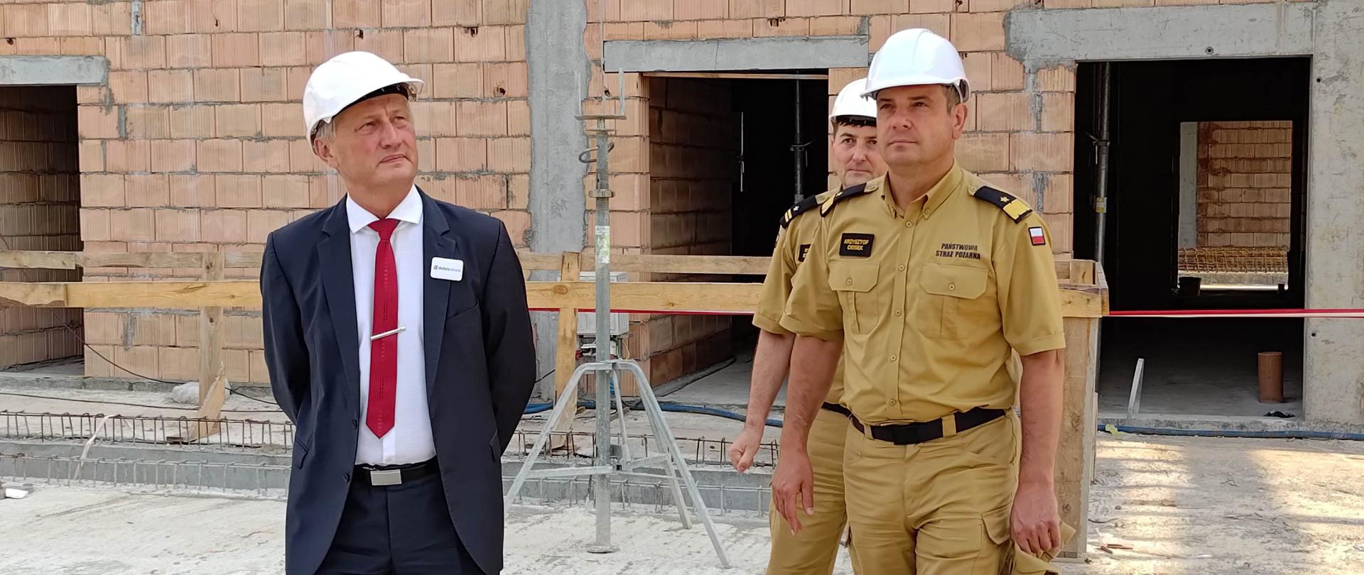 Na zdjęciu widzimy dwóch mężczyzn w mundurach PSP oraz mężczyznę w garniturze podczas doglądania budowy strażnicy w Końskich. Wszyscy Panowie mają kaski białe na głowach.