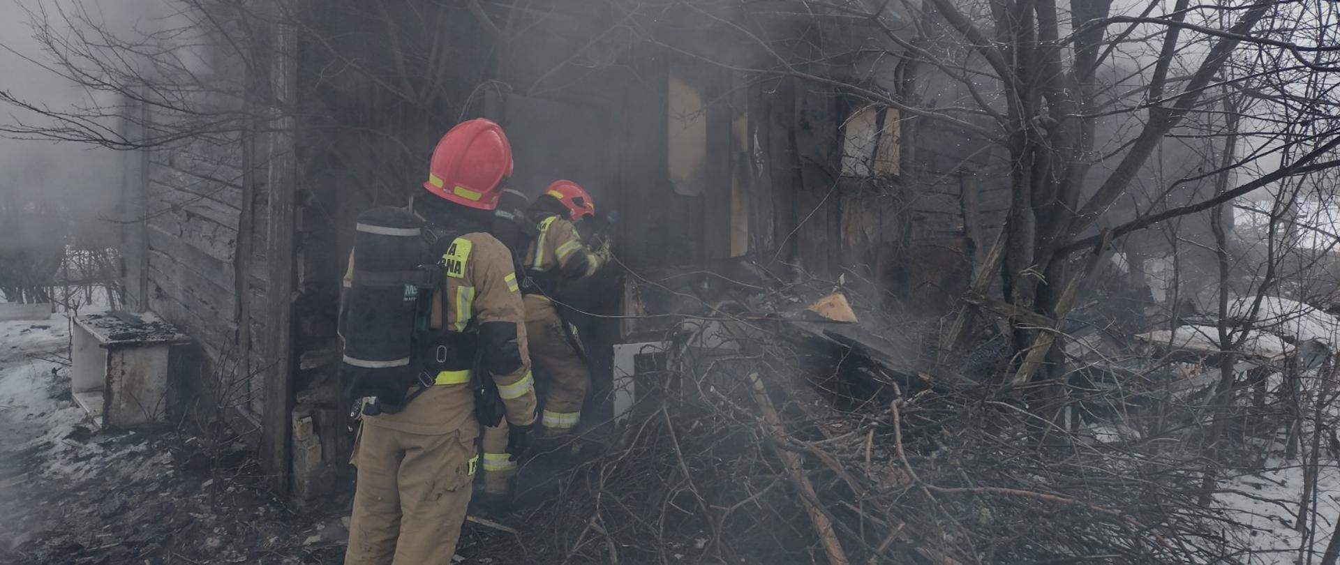 Na zdjęciu w porze dziennej widać dwóch strażaków przed budynkiem drewnianym, którzy gaszą wodą pożar. Na zdjęciu panuje silne zadymienie na zewnątrz.