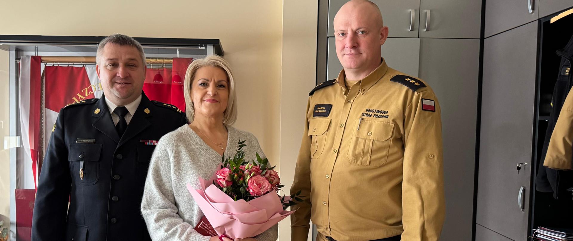 Na pierwszym planie widoczni są: Komendant Powiatowy Państwowej Straży Pożarnej w Rypinie st. bryg. Dariusz Łęgosz, Główna Księgowa Joanna Marchlewska oraz Zastępca Komendanta Powiatowego PSP kpt. Marcin Czarnecki.
Główna księgowa trzyma w dłoni otrzymany list gratulacyjny oraz kwiaty, a w tle widoczny jest sztandar Państwowej Straży Pożarnej.