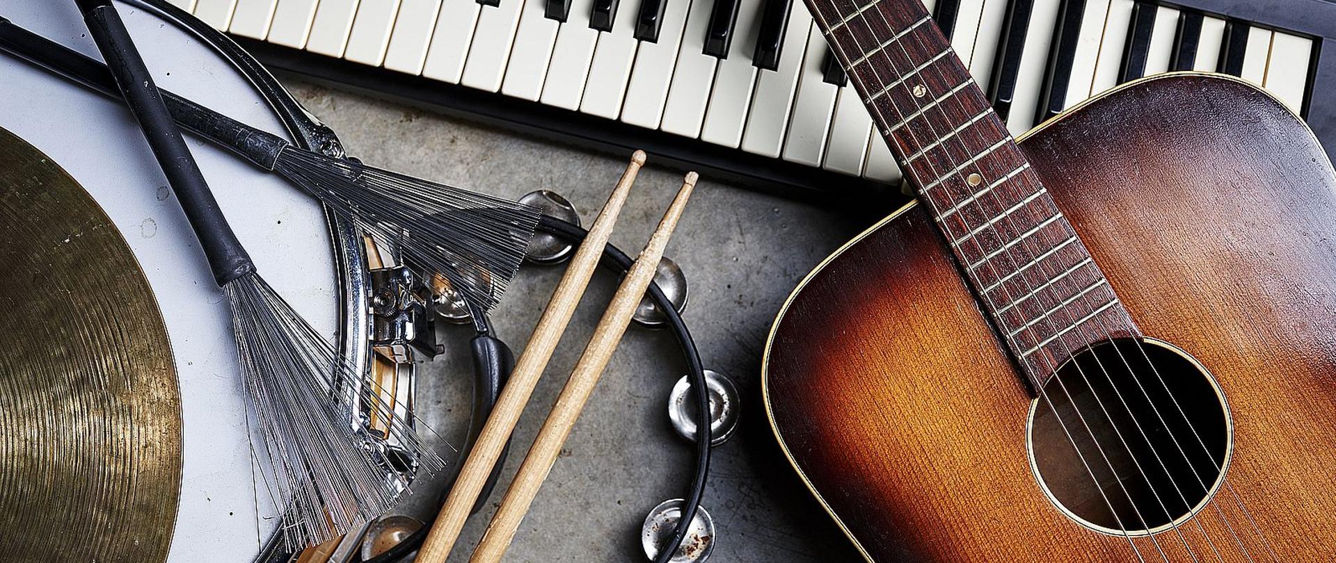 Zdjęcie przedstawia klawiaturę pianina, gitarę oraz instrumenty perkusyjne jak werbel, tamburyn i talerz perkusyjny leżące na podłodze.