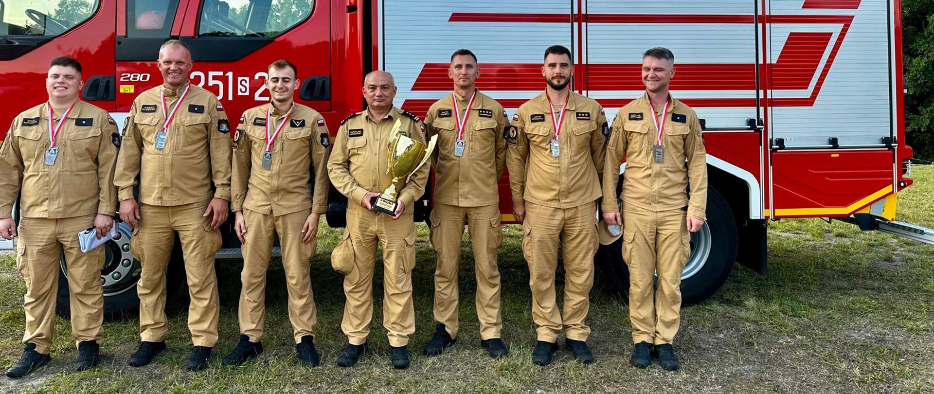 Pamiątkowe zdjęcie grupowe na tle pożarniczego samochodu bojowego na którym stoi Komendant Centralnej Szkoły PSP trzymający w dłoni puchar w towarzystwie strażaków drużyny CS PSP która zajęła drugie miejsce w zawodach. Zawodnicy maja na szyi zawieszony medal
