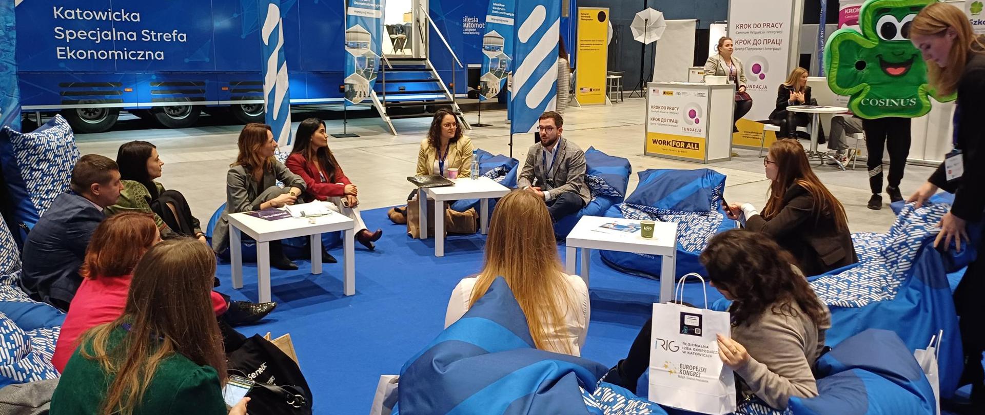 Grupa osób siedzi na niebieskich pufach w okręgu. Z tyłu widać samochód z napisem Katowicka Specjalna Strefa Ekonomiczna