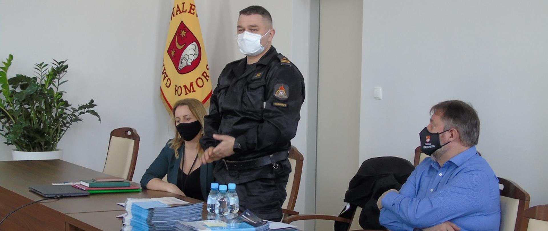 NA ZDJĘCIU WIDAC 2 MĘŻCZYZN I KOBIETĘ. MĘŻCZYZNA W ŚRODKU STOI , POZOSTALI SIEDZĄ PRZY STOLE NA KÓRYM LEŻY KILKA STOSÓW ULOTEK, JEDEN Z NICH MA W DŁONI ULOTKI .