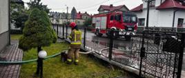 Na zdjęciu widzimy strażaka obsługującego motopompę podczas działań ratowniczych polegających na pompowaniu wody z zalanej piwnicy. W tle pojazd pożarniczy. 