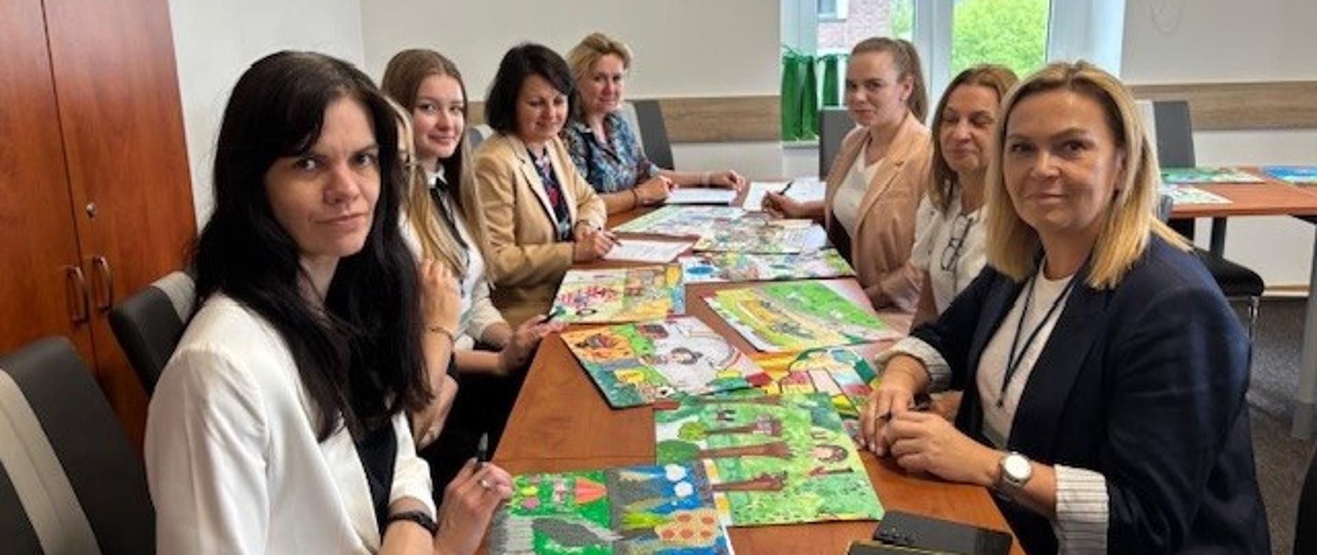 Osiem kobiet siedzi po dwóch stronach stołu, na którym leżą kolorowe rysunki dzieci. Kobiety uśmiechają się pozując do zdjęcia.