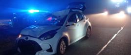 Biały samochód osobowy stoi na drodze, ma rozbity cały przód i otwartą klapę bagażnika. Za nim stoi radiowóz policyjny o raz zastęp PSP . Pora nocna. 
