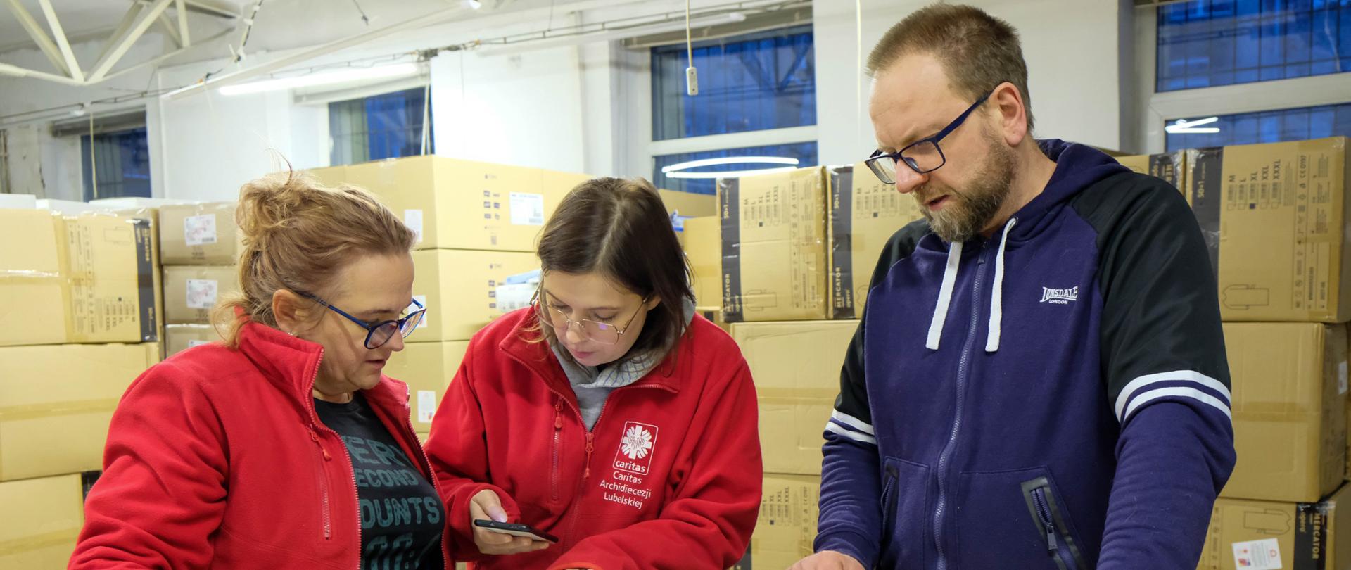 W magazynie pełnym paczek z pomocą materialną obok jednego stosu pudeł stoją dwie kobiety i mężczyzna i patrzą się na leżące na pudłach kartki