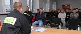 strażak w ubrania koszarowym państwowej straży pożarnej siedzi przodem przy stoliku do zebranych na sali kilkunastu osób ubranych w ubrania koszarowe ochotniczej straży pożarnej barwy czarnej oraz ubrania cywilne siedzi na krzesełkach i słucha przemówienia zastępcy dowódcy Jednostki Ratowniczo-Gaśniczej w Łowiczu w tle wisząca szafka po lewej stronie widoczne cztery okna w prawym rogu widoczne dwa puste krzesła z czarnym materiałowym obiciem na podłodze panele barwy jasnoszarej