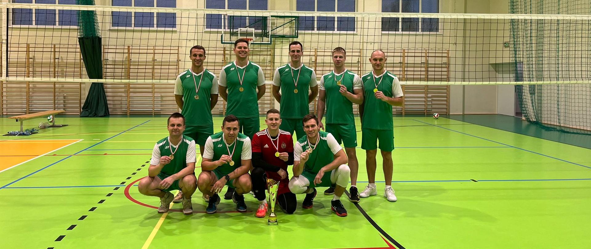 Strażacy stoją na sali sportowej w zielonych strojach i medalami i pucharem