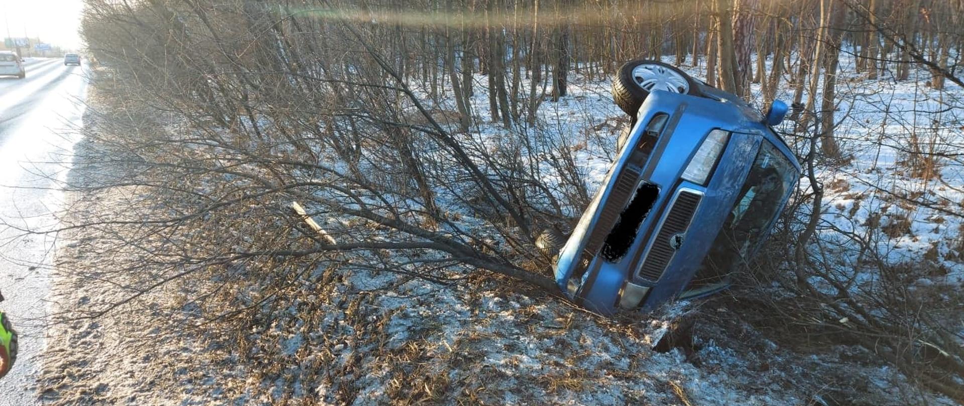 Pojazd marki Skoda koloru niebieskiego leżący na lewym boku na poboczu drogi, sceneria zimowa
