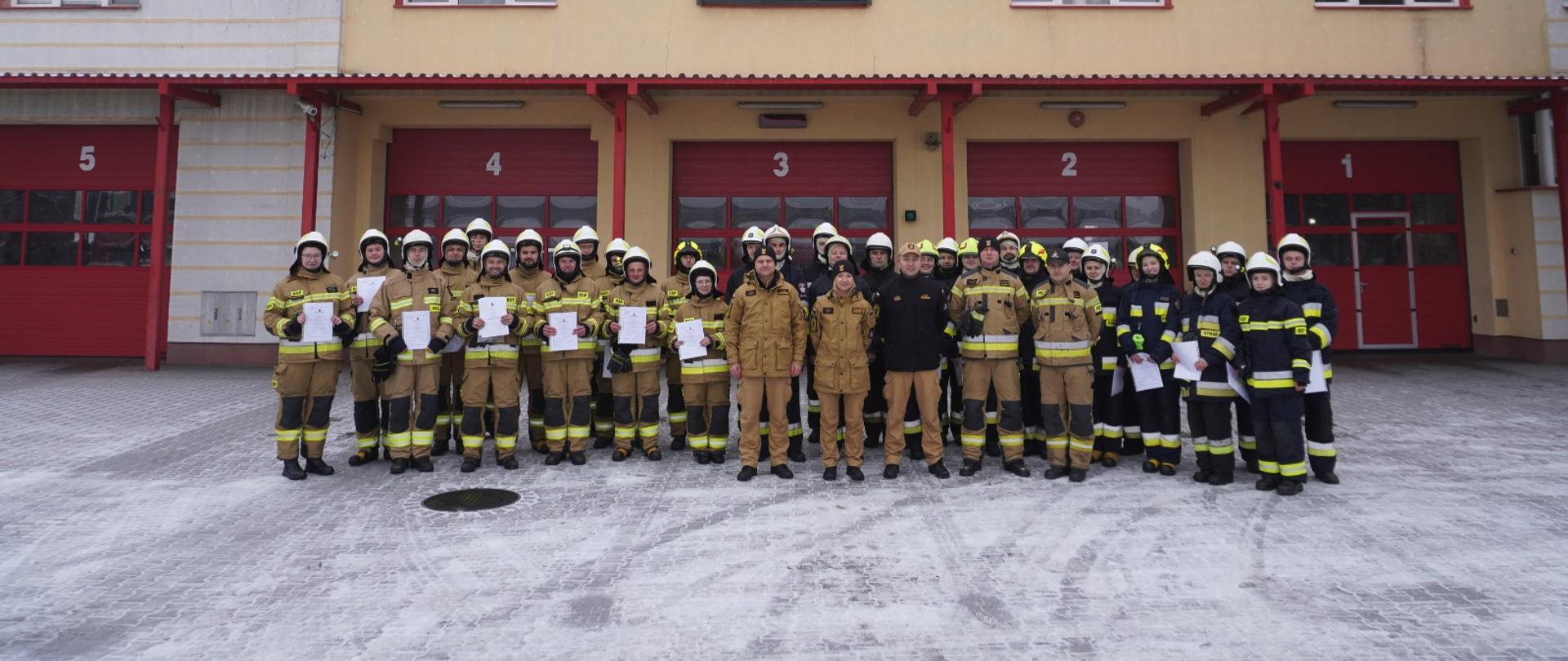 Pochmurny zimowy dzień na tle budynku Komendy Powiatowej Państwowej Straży Pożarnej w Chojnicach do zdjęcia pozuje grupa strażaków ubranych w piaskowe i czarne ubrania specjalne prezentując dyplomy ukończenia szkolenia podstawowego Strażaka Ratownika OSP. 