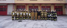 Pochmurny zimowy dzień na tle budynku Komendy Powiatowej Państwowej Straży Pożarnej w Chojnicach do zdjęcia pozuje grupa strażaków ubranych w piaskowe i czarne ubrania specjalne prezentując dyplomy ukończenia szkolenia podstawowego Strażaka Ratownika OSP. 