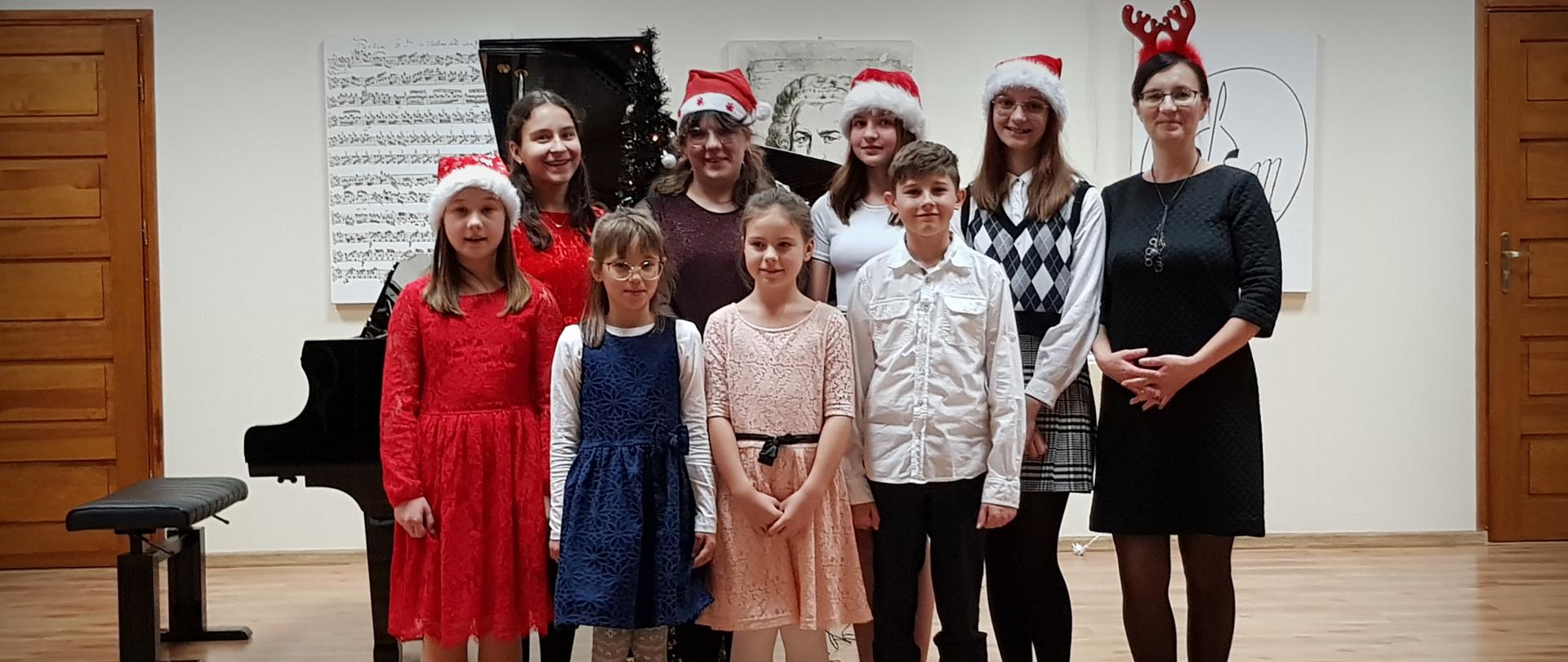 Uczniowie klasy fortepianu stoją wraz z nauczycielem na tle fortepianu. Uczniowie mają na głowach czapki Mikołaja.