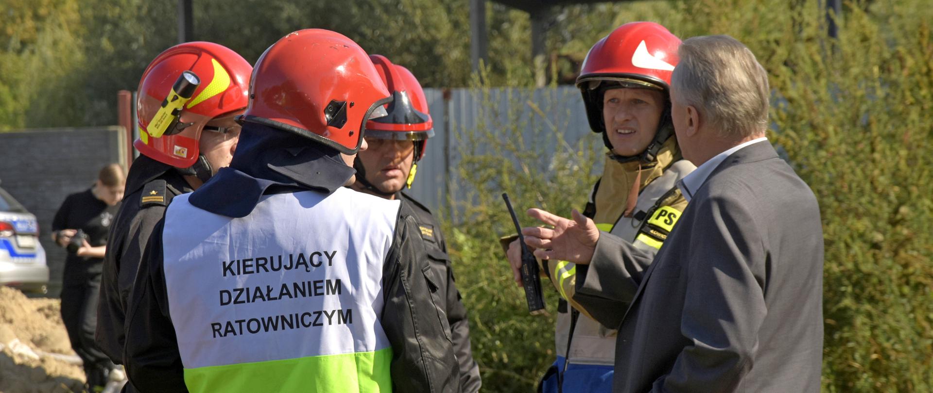 czterech strażaków rozmawia z osobą cywilna ubrana w garnitur, która ma uniesioną prawa dłoń lekko do góry, strażacy maja hełmy na głowie, strażak na pierwszy planie ma założoną kamizelkę z napisem „kierujący działaniem ratowniczym”, w tle widać zarośla, na dalszy planie metalowy płot z blachy przed którym stoi samochód osobowy i osoba z aparatem fotograficznym w dłoniach, obok tej osoby kupka piachu. Pogoda słoneczna.