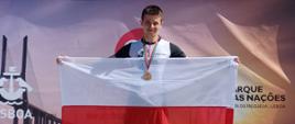 Zdjęcie wykonano w ciągu dnia, na podium stoi ubrany w kombinezon triathlonowy Łukasz Krieger w rękach trzymając polską flagę pozuje do zdjęcia. Na szyi ma zawieszony złoty medal. W tle banery reklamowe promujące wydarzenie sportowe. 