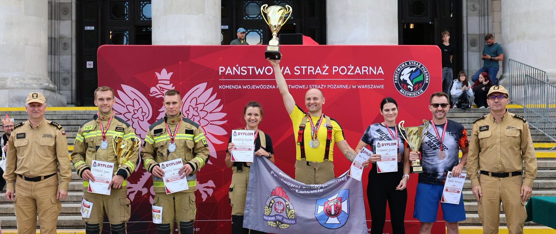 Laureaci zawodów wraz z przedstawicielami Państwowej Straży Pożarnej pozują przed czerwoną ścianką PSP przy Pałacu Kultury i Nauki. Zawodnicy prezentują puchary, medale, dyplomy oraz flagę swojej jednostki.