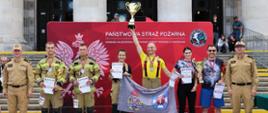 Laureaci zawodów wraz z przedstawicielami Państwowej Straży Pożarnej pozują przed czerwoną ścianką PSP przy Pałacu Kultury i Nauki. Zawodnicy prezentują puchary, medale, dyplomy oraz flagę swojej jednostki.