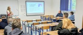 Uczniowie siedzący w ławkach słuchają prelekcji prowadzonej przez kobietę. Na tablicy wyświetlona jest prezentacja.