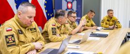 Pięciu oficerów PSP uczestniczy w obradach zespołu w sali konferencyjnej. Każdy pracuje na laptopie lub robi notatki. W tle widoczne flagi Polski i Unii Europejskiej oraz baner Komendy Głównej PSP.