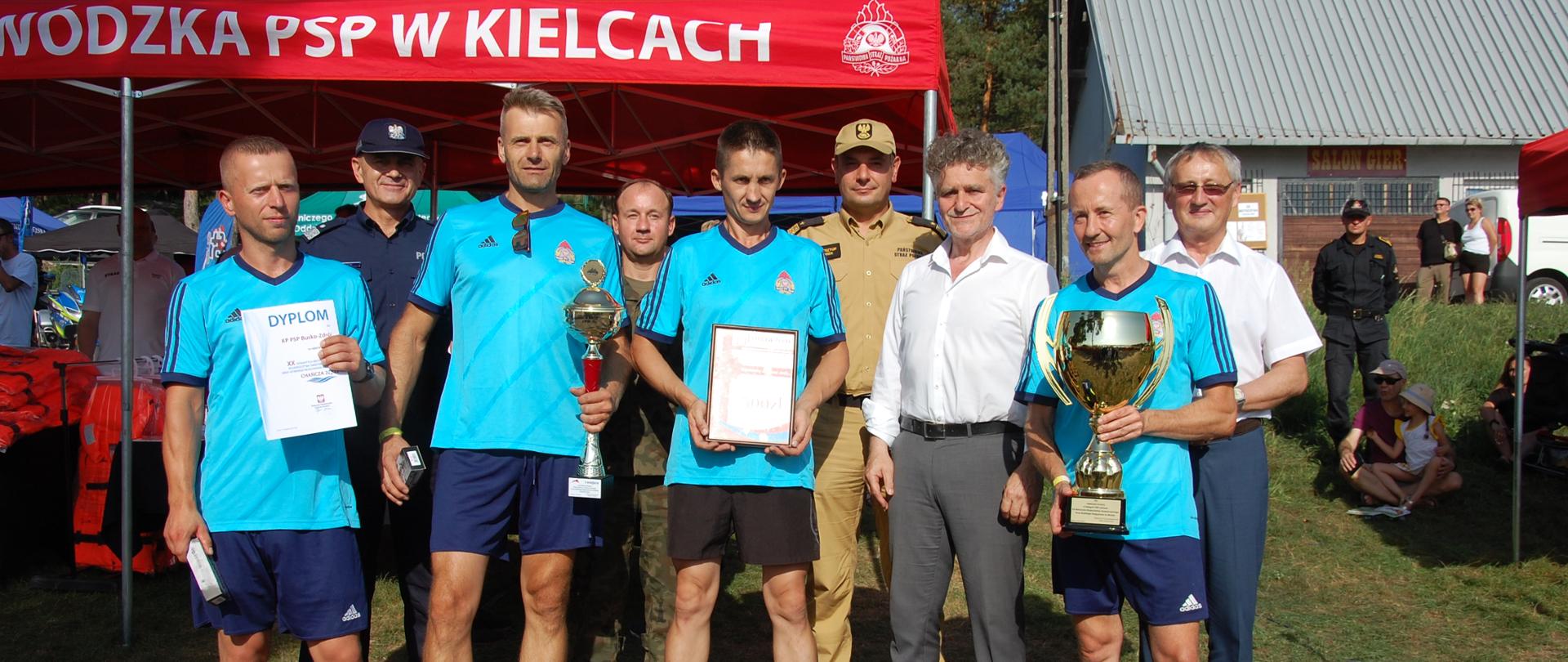 Na zdjęciu trzech mężczyzn w mundurach w drugim rzędzie oraz dwóch w białych koszulach stoją za czteroma mężczyznami ubranymi w stroje sportowe z krótkimi rękawami i nogawkami koloru niebieskiego. Sportowcy trzymają w rękach nagrody.
