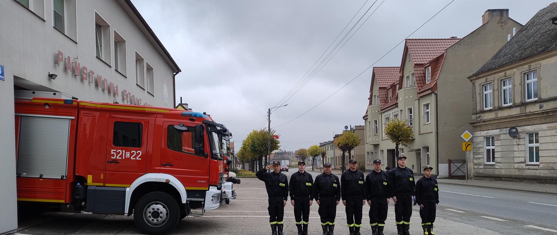 strażacy w ubraniach koszarowych stoją w szeregu przed budynkiem Komendy Powiatowej Państwowej Straży Pożarnej w Sępólnie Krajeńskim w tle stoi średni samochód ratowniczo-gaśniczy.