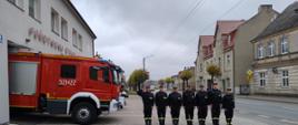 strażacy w ubraniach koszarowych stoją w szeregu przed budynkiem Komendy Powiatowej Państwowej Straży Pożarnej w Sępólnie Krajeńskim w tle stoi średni samochód ratowniczo-gaśniczy.