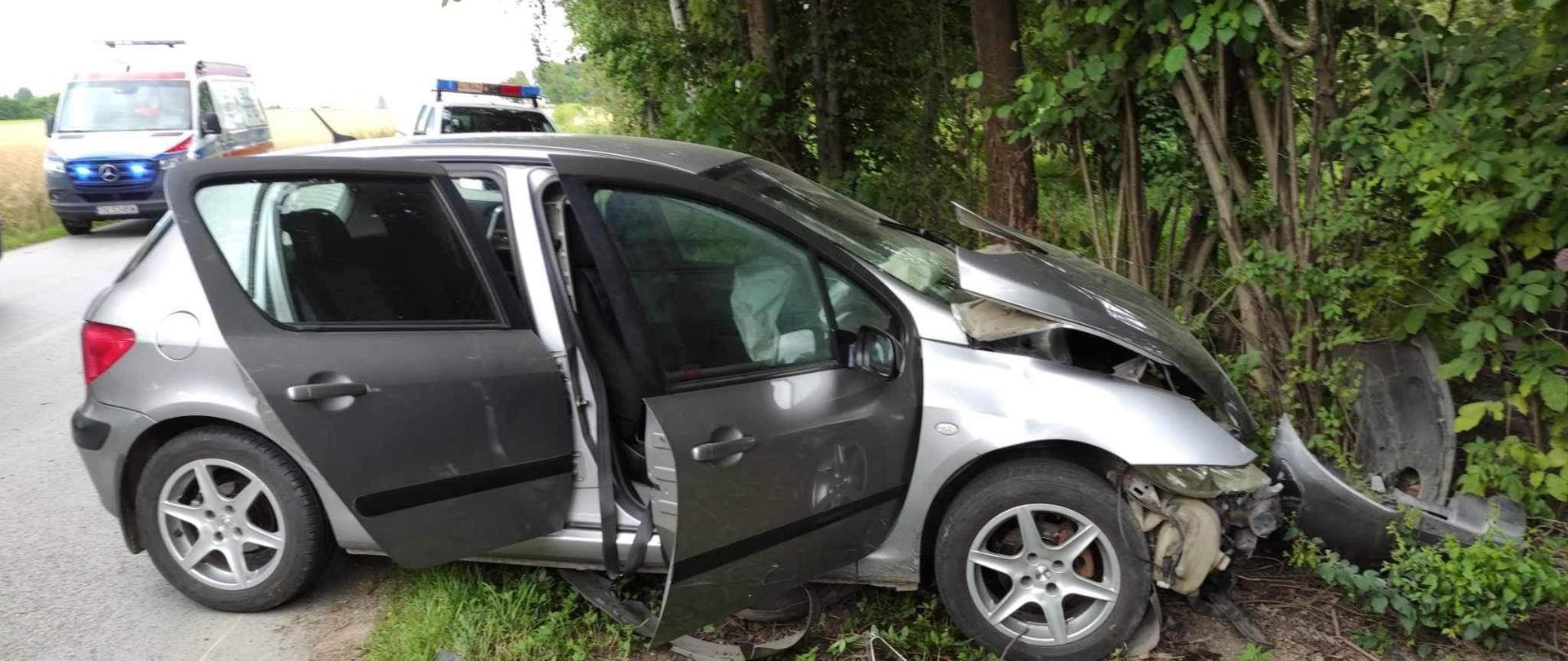 Na zdjęciu widoczny jest samochód osobowy z uszkodzonym przodem w wyniku zdarzenia drogowego. samochód znajduje się na poboczu drogi. W tle widoczna jest karetka pogotowia oraz radiowóz policji.