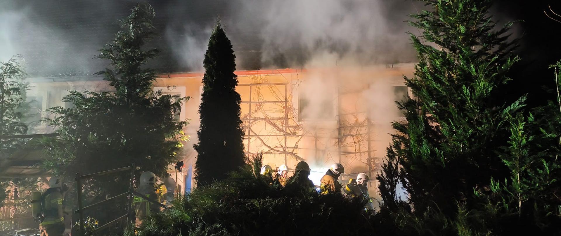 budynek mieszkalny w tle, widać dym, noc, strażacy w tle i drzewa