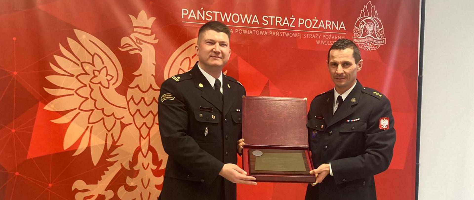 Zdjęcie wykonane wewnątrz pomieszczenia. Przedstawia Z-ce komendanta Powiatowego PSP w Wolsztynie wraz z Komendantem Powiatowym PSP w Wolsztynie. W tle baner w kolorze czerwonym z napisem Państwowa Straż Pożarna.