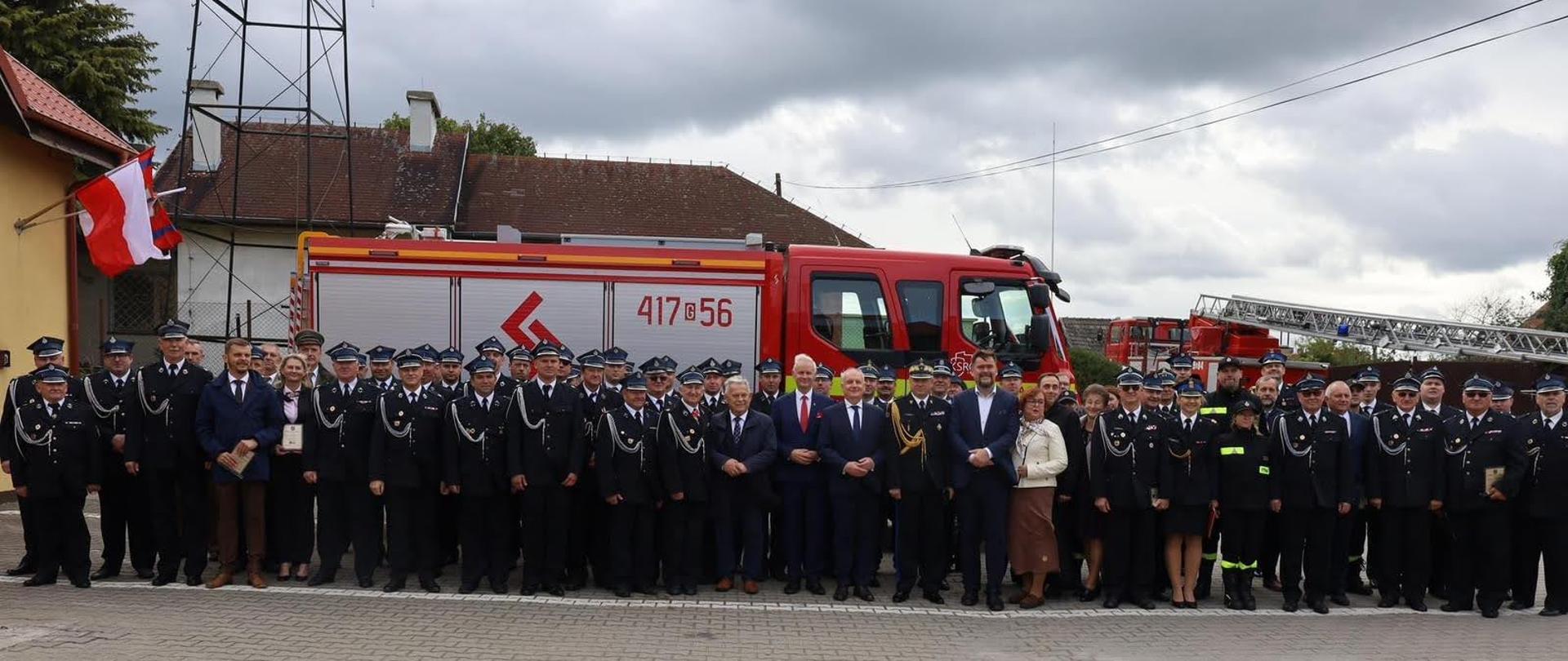 Strażacy ochotniczej straży pożarnej funkcjonariusze Państwowej Straży Pożarnej oraz zaproszeni gości stoją obok siebie za nimi stoi samochód gaśniczy oraz samochód specjalny drabina obok na budynku jednostki wiszą na ścianie flagi Polski.