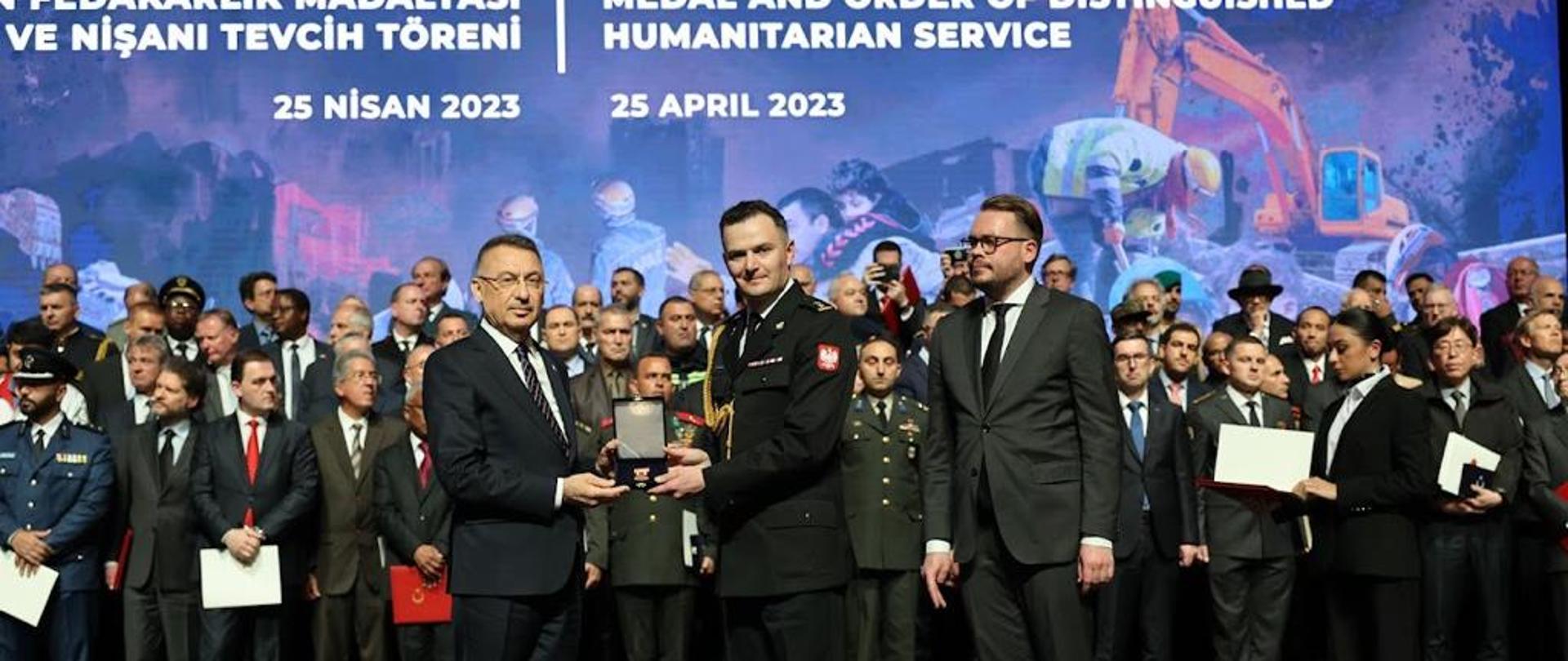 Bryg. Borowiec stoi na scenie wspólnie z przedstawicielem Turcji , obaj wspólnie podtrzymują etui z orderem, obok nich stoi po prawej stronie mężczyzna w garniturze, w tle grupa innych uczestników